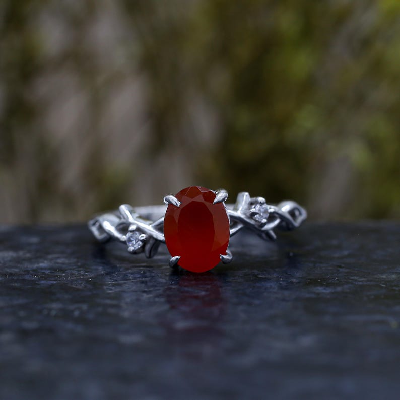 May include: A silver ring featuring a central oval-cut deep orange carnelian stone held by four prongs. The band is designed with a textured, vine-like motif and includes two small, clear, round-cut accent stones set on either side of the center.