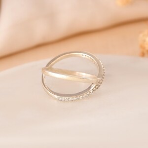 Puede incluir: Un anillo plateado con un dise&ntilde;o &uacute;nico. Una banda cruza otra, con peque&ntilde;as piedras transparentes engastadas a lo largo de un lado de la banda superior. El anillo se muestra sobre una superficie blanca con un fondo suave y neutro.