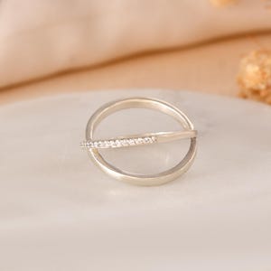 Puede incluir: Un anillo de plata con un dise&ntilde;o &uacute;nico. El anillo presenta una banda circular cruzada por una barra recta adornada con peque&ntilde;as piedras brillantes. El anillo se encuentra sobre un fondo suave y neutro, resaltando su elegancia.
