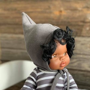 Könnte beinhalten: Ein graues und weiß gestreiftes Baby-Puppen-Schlafanzug mit einer grauen Kapuze. Die Puppe hat braune Augen, dunkelbraunes lockiges Haar und braune Haut.