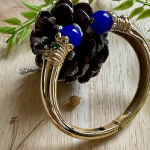 May include: Gold-coloured hinged bangle bracelet with blue cabochon accents and matching earrings. The earrings feature a bow design, green emerald-cut stones, and blue cabochon drops. The jewellery is displayed on a wooden surface.