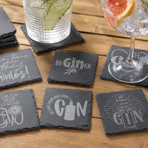Könnte beinhalten: Ein Set quadratischer Untersetzer aus dunkelgrauem Schiefer mit rauen, natürlichen Kanten. Jeder Untersetzer ist mit einzigartigen, weißen Lasergravuren versehen, die Texte und grafische Motive rund um das Thema Gin zeigen.