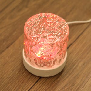 Puede incluir: Una pequeña lámpara redonda de cristal rosa con una superficie texturizada. La lámpara se asienta sobre una base blanca y emite un brillo rosa cálido. Un cable blanco se extiende desde la lámpara, lo que sugiere que funciona con electricidad. La lámpara está sobre una superficie de madera.