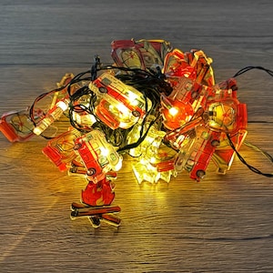 Puede incluir: Cadena de luces decorativas con mini furgonetas rojas y amarillas y un diseño de fogata. Las luces están en un cable negro y emiten un brillo cálido. Ideal para añadir un toque de diversión a cualquier espacio.