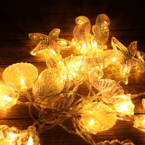 May include: String of warm white LED lights shaped like seashells, seahorses, and starfish. The lights are strung on a clear wire and are illuminated, creating a soft, ambient glow. The decorative lights are suitable for indoor or outdoor use.