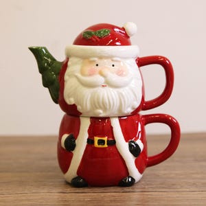May include: A festive red and white ceramic teapot and teacup set shaped like Santa Claus. The teapot features a green tree-shaped spout and a red hat with holly. The teacup is the base of Santa's body, with a gold belt buckle.