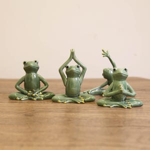 Puede incluir: Cuatro figuras de ranas de cerámica verde en posturas de yoga. Las ranas están en varias posiciones, incluida la postura del loto con las manos en oración y los brazos levantados. Las figuras están sobre una superficie de madera.
