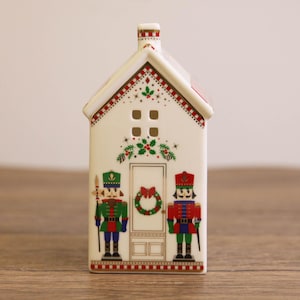 Puede incluir: Una casa navideña de cerámica blanca con techo y chimenea rojos. La casa presenta una puerta con una corona y dos soldados cascanueces. La casa está decorada con detalles rojos, verdes y dorados, ideal para la decoración navideña.