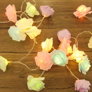 May include: A decorative string of LED lights featuring pastel-coloured rose-shaped ornaments. The roses are in shades of pink, purple, and white, with a warm yellow glow. The string is arranged on a wooden surface.