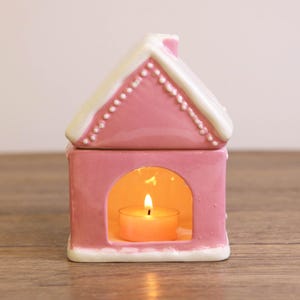 Può includere: Un portacandele in ceramica rosa a forma di casa, con tetto e finiture bianche. La casa ha una base rettangolare con un'apertura ad arco e una candela accesa all'interno. Il tetto è decorato con dettagli bianchi.