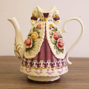 Puede incluir: Una tetera decorativa de porcelana con forma de vestido. La tetera presenta un corpiño color crema con detalles morados y está adornada con rosas rojas y amarillas. El asa y el pico son de color crema con detalles dorados.