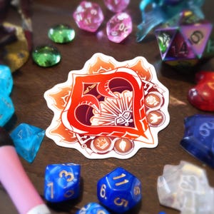 Peut inclure: Un autocollant en forme de cœur aux tons rouges et orangés, orné d'éléments décoratifs. L'image présente également divers dés polyédriques colorés, dont des dés bleus, roses et transparents, disposés sur une surface en bois.