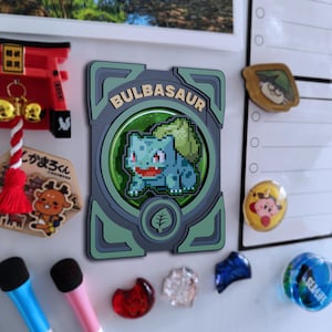 May include: A Bulbasaur magnet with a pixelated design. The magnet features a green and blue Bulbasaur character inside a green and grey frame. The word "Bulbasaur" is printed at the top of the frame.
