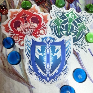 Puede incluir: Tres pegatinas decorativas con diseños intrincados. Una es roja con forma de corazón, otra verde con un patrón circular y la tercera azul con forma de escudo. Las pegatinas están rodeadas de cuentas de vidrio azules y verdes y dados.