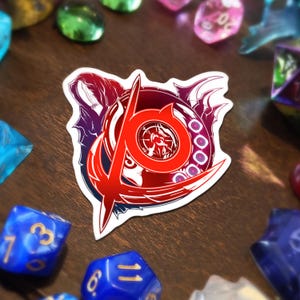 Può includere: Un adesivo con un design grafico rosso e bianco, con una lettera "A" stilizzata e un emblema circolare. L'adesivo ha un bordo bianco ed è circondato da dadi colorati su una superficie di legno.