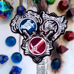 Può includere: Tre adesivi in bianco e nero con disegni circolari raffiguranti una spada, uno scudo e un teschio. Vari dadi poliedrici blu, rossi e verdi e sfere di vetro blu circondano gli adesivi. Tema fantasy.