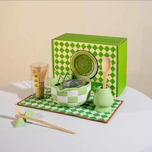 May include: A matcha tea set featuring a green and white chequered bowl, a bamboo whisk, a wooden spoon, and a green ceramic spoon holder. The set includes a green box with a matcha tea image and a chequered pattern.
