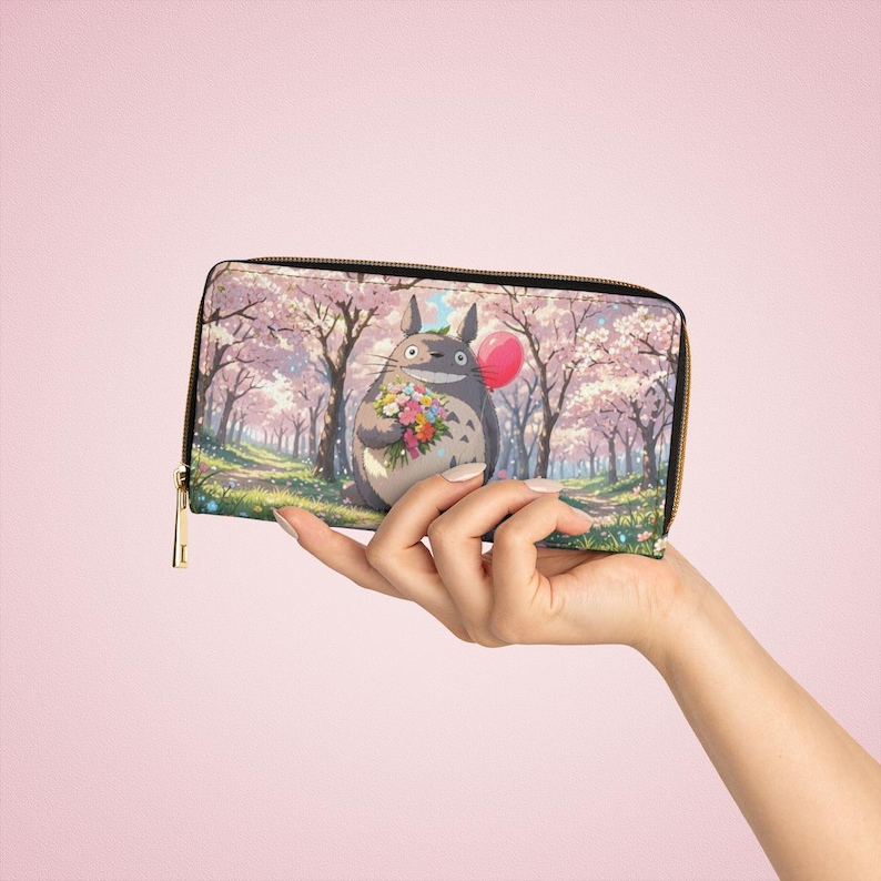 May include: A zip-around wallet with a colourful illustration of a Totoro character holding flowers and a balloon, set against a cherry blossom forest. The wallet has a black zip and a gold-coloured zip pull.