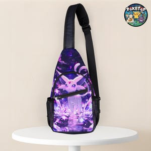 Puede incluir: Bolso bandolera morado y negro con un diseño de criatura fantástica. El bolso tiene una correa ajustable negra y un compartimento principal con cremallera. El diseño incluye una criatura morada con un fondo estrellado y detalles de cristal. El bolso está sobre una mesa blanca.