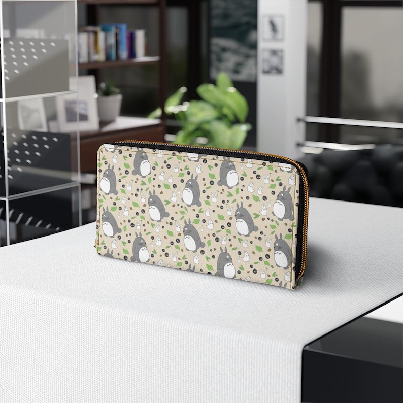 May include: Tan zip-around wallet with a repeating pattern of grey Totoro characters, white rabbit-like creatures, green leaves, and black dots. The wallet has a gold-coloured zip and a beige interior. The wallet is on a white surface.