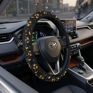 May include: Black steering wheel cover with a repeating pattern of yellow cartoon faces, stars, and crescent moons. The cover is installed on a black steering wheel inside a car. The car's dashboard and infotainment screen are visible in the background.