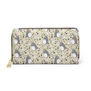May include: A beige wallet featuring a repeating pattern of grey Totoro characters, white sprites, green leaves, and black berries. The wallet has a gold zip and pull.