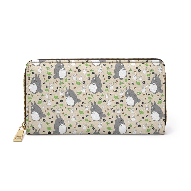 May include: A beige wallet featuring a repeating pattern of grey Totoro characters, white sprites, green leaves, and black berries. The wallet has a gold zip and pull.