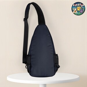 May include: Navy blue sling bag with a black adjustable strap and a black mesh pocket on each side. The bag has a rounded shape and a black padded shoulder strap. A circular logo with cartoon characters is visible in the upper right corner.