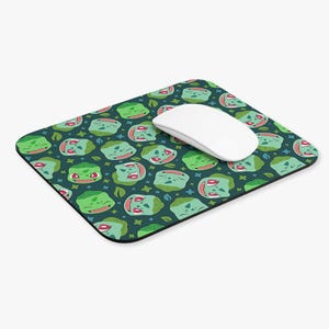 May include: A rectangular mousepad featuring a repeating pattern of green cartoon character faces on a dark teal background. A white wireless mouse rests on the mousepad. The mousepad has a black border.