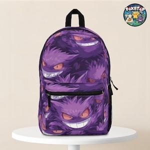 May include: Purple backpack featuring a repeating pattern of smiling cartoon characters with sharp teeth and red eyes. The backpack has a black handle and zipper, and a side pocket. A circular logo with cartoon characters is visible.