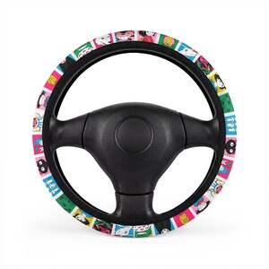 May include: A black steering wheel cover featuring a repeating pattern of colorful square tiles. Each tile displays a different cartoon character illustration in various colors including pink, green, blue, and yellow against a white background.