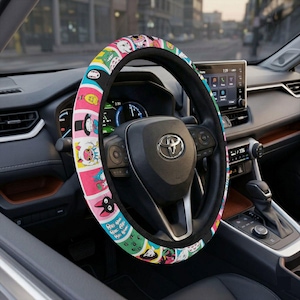 May include: A colorful steering wheel cover featuring a cartoon character design. The cover is primarily pink, blue, and green, with a black border. The cover is installed on a black steering wheel inside a car.