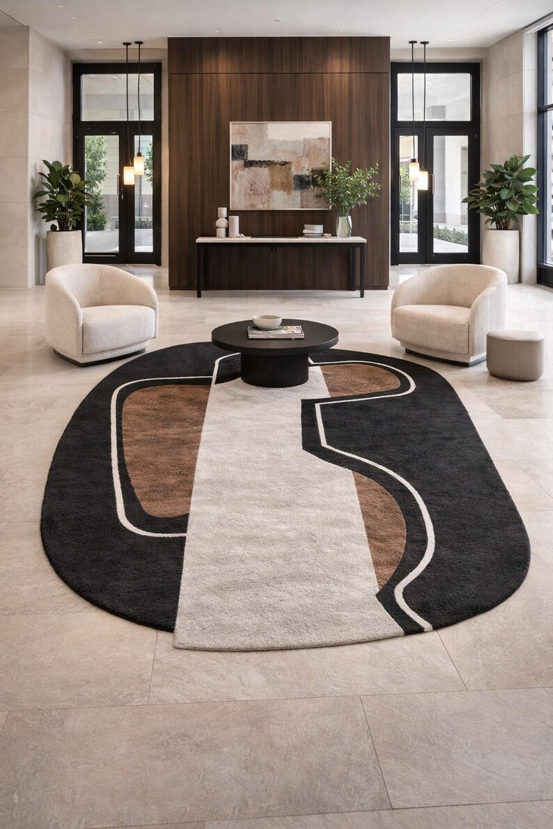 May include: An oval area rug with a geometric design in black, brown, and cream colors. The rug is placed in a modern living space with two cream-colored armchairs and a dark brown coffee table.