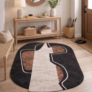 May include: Oval area rug with a modern design. The rug features a black background with a white, brown, and beige geometric pattern. The rug is placed on a hardwood floor in a well-lit entryway.