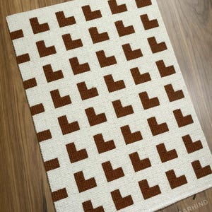 May include: A rectangular, off-white rug with a repeating pattern of brown L-shaped designs. The rug is made of a textured material and is placed on a wooden floor. The design is consistent across the rug's surface.