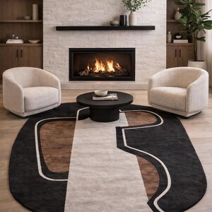 May include: An oval area rug with a geometric design in black, brown, and cream colors. The rug is placed in front of a fireplace with two cream-colored armchairs and a black coffee table.