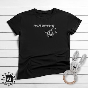 Op de afbeelding: Zwart T-shirt met de tekst "not AI generated" en een grillige krabbel van een karakter. Een wit, gehaakt konijnenspeeltje met een houten ring ligt naast het shirt. Het shirt ligt op een wit houten oppervlak.