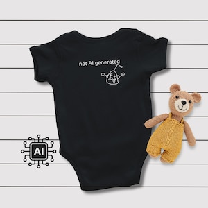 May include: A black short-sleeved baby bodysuit featuring the white text "not AI generated" alongside a simple white line drawing of a robot. A small brown crocheted teddy bear wearing yellow overalls is positioned next to the bodysuit.