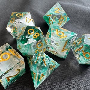 Op de afbeelding: Set van zeven polyhedrale dobbelstenen met een heldere harsbasis, gemengd met groen en wit. De cijfers zijn goudkleurig. De dobbelstenen zijn ontworpen voor tabletop gaming.