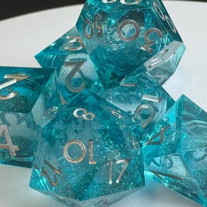 May include: A set of seven polyhedral dice in shades of teal and clear resin. The dice have silver numbering and internal glitter. The dice are designed for tabletop role-playing games.