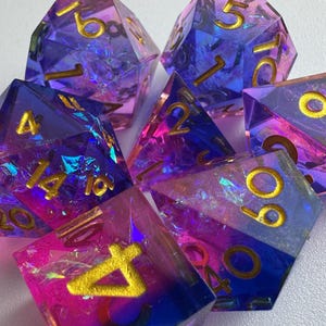Puede incluir: Un conjunto de siete dados poliédricos en tonos de azul, morado y rosa con numeración dorada. Los dados están hechos de una resina transparente con inclusiones iridiscentes, creando un efecto brillante. Los dados son para juegos de mesa.