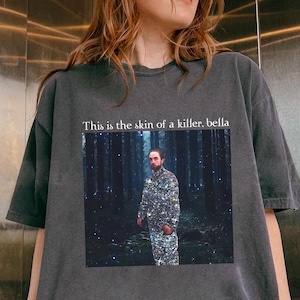 May include: Dark gray t-shirt featuring a graphic of a man in a sparkly suit standing in a forest. The text "This is the skin of a killer, bella" is printed above the graphic. The shirt is worn by a person.