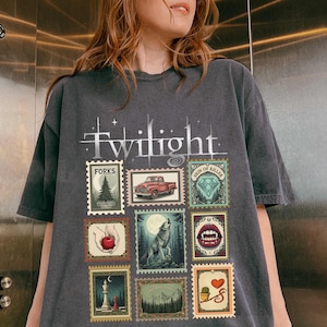 May include: Dark grey t-shirt featuring the word "Twilight" and several postage stamp-style graphics. The stamps depict a red truck, a wolf, a forest, and other related imagery. The shirt has a vintage, worn appearance.