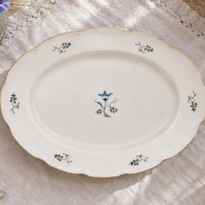 May include: An oval white ceramic serving platter with a scalloped edge and a thin gold rim. The center features a single blue flower illustration, while the perimeter is decorated with six small, delicate black floral sprig motifs.