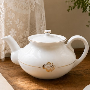 Pode incluir: Bule de cerâmica branco com acabamento dourado e design floral. O bule tem uma alça curva e um bico. Uma tampa combinando fica no topo. Em uma superfície de madeira, com uma xícara e pires em primeiro plano.
