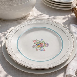 May include: A white porcelain plate with a floral pattern and a light blue border. The plate is part of a set, with a soup tureen and other plates visible. A silver spoon and a basket of bread are also present.