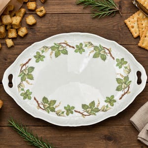 Op de afbeelding: Een ovale, witte serveerschaal met een decoratieve rand van groene bladeren en wijnranken. De schaal heeft twee handvatten en is omgeven door crackers, breadsticks, croutons en rozemarijntakjes op een houten oppervlak.