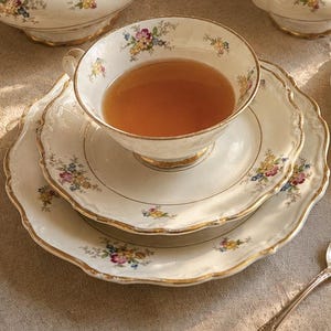 Pode incluir: Um conjunto de chá em porcelana com base branca, decorado com delicados padrões florais em tons de rosa, amarelo e azul, com bordas douradas. O conjunto inclui duas chávenas com pires, um açucareiro com tampa, uma leiteira e uma colher.