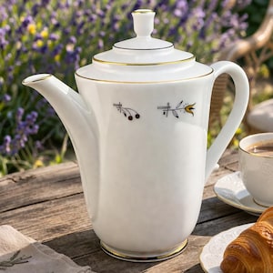 Pode incluir: Uma cafeteira de porcelana branca com detalhes dourados, uma chávena e pires a condizer e um croissant num prato estão numa mesa de madeira. Uma guardanapo de linho dobrado com um desenho de raminho também está presente.