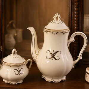 May include: A white porcelain teapot and matching sugar bowl with a scalloped base and rim. Both pieces feature gold-toned trim and a central dark brown stylized X-shaped monogram. The teapot has a curved handle and spout with a lid.